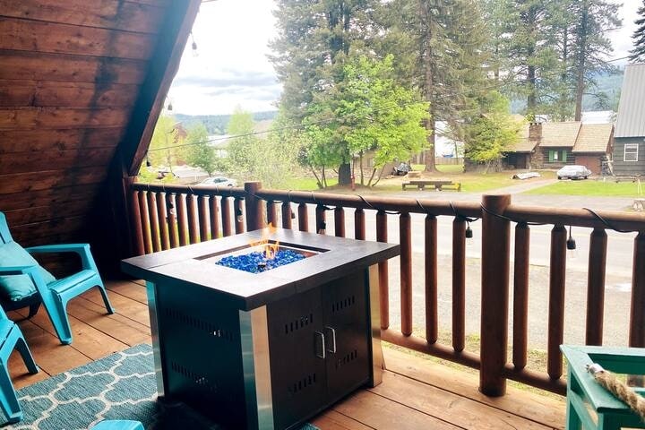 The outdoor seating area features a contemporary fire table, surrounded by blue chairs. A scenic view of the trees and lake is visible beyond the railing. Soft lighting from hanging bulbs adds to the inviting environment.