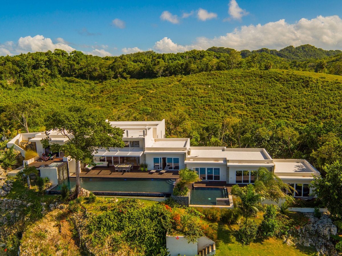 A modern villa is presented from an aerial perspective, showcasing its elongated structure and clean lines. The surrounding lush greenery complements the property, while the infinity pool reflects the sky. Large glass windows invite light and provide views of the natural landscape.