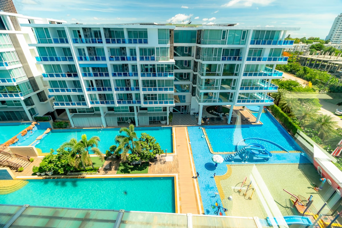 A view from above showcases the resort's extensive pool area, featuring multiple bright blue swimming pools surrounded by well-maintained greenery. Sun loungers are positioned near the water, and a children's playground is visible, enhancing the family-friendly atmosphere.
