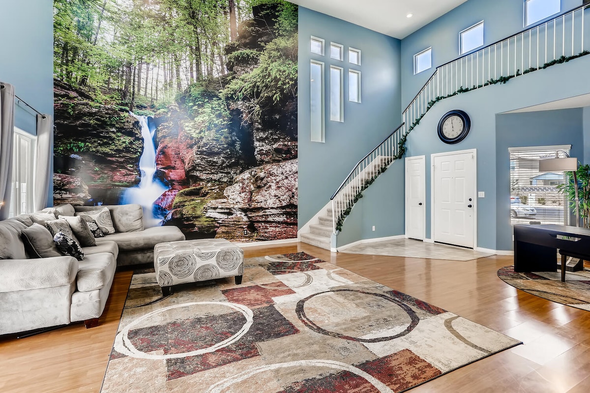 A spacious living area is highlighted by a large mural of a waterfall set against lush greenery. Light-colored furniture is arranged around a circular patterned rug, while tall windows allow natural light to fill the room. A staircase leads to the upper level.