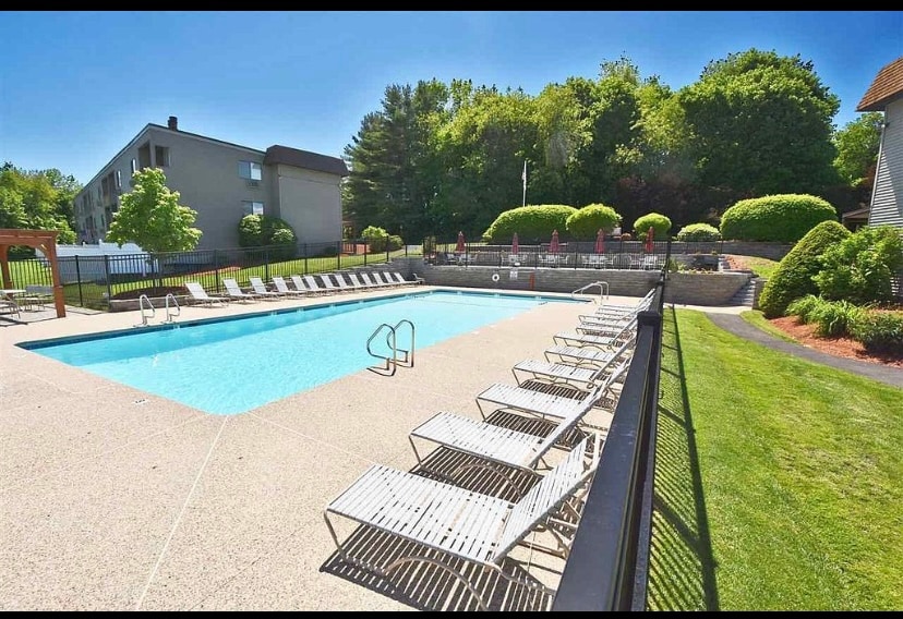 A spacious saltwater pool area is surrounded by lounge chairs arranged neatly along the poolside. Lush greenery and hedges provide a natural backdrop, while a fenced perimeter ensures privacy. Wooden seating areas are visible in the distance, creating an inviting outdoor space.