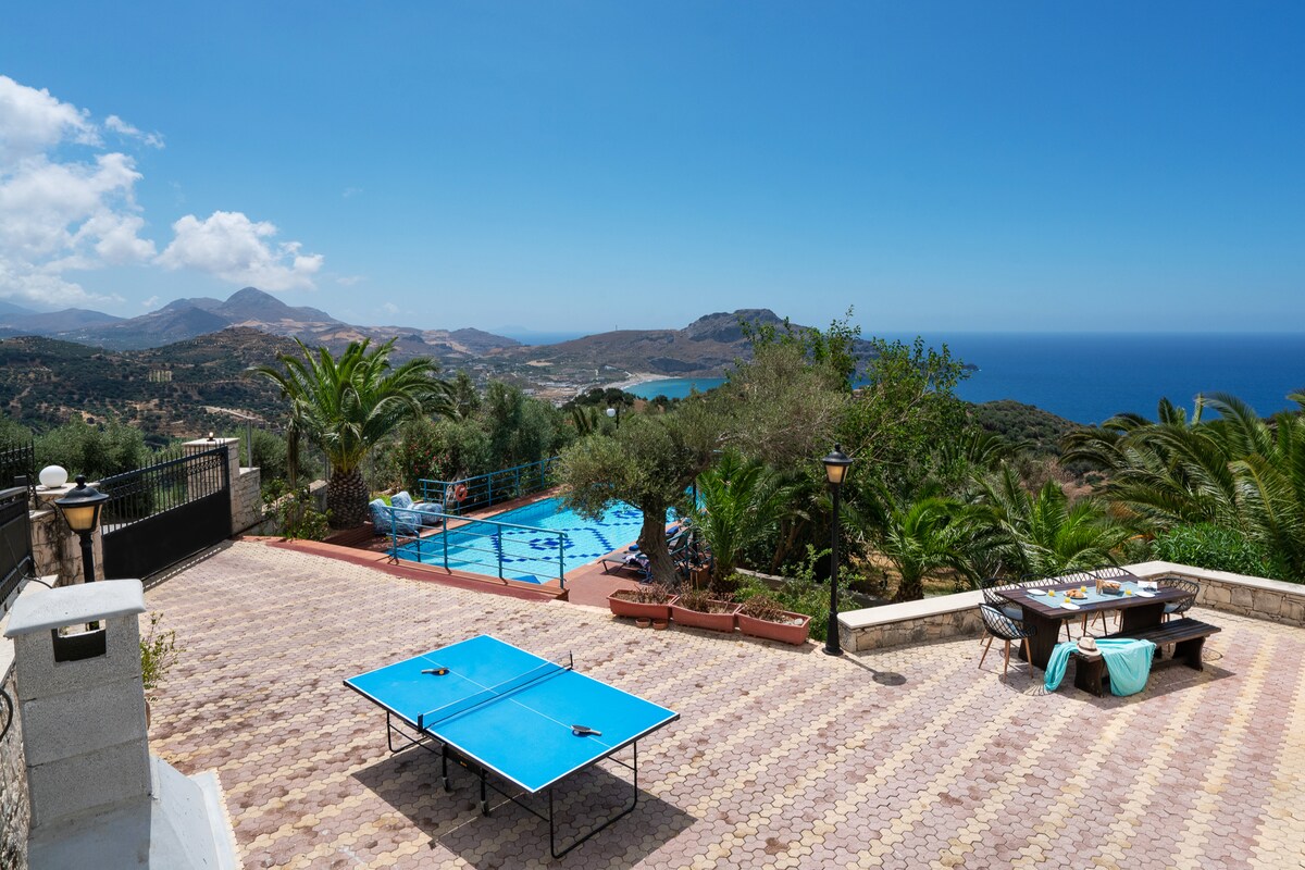 An outdoor area features a private swimming pool surrounded by sunbeds and puffs. A ping pong table is positioned on the terrace, with lush greenery and panoramic views of the Libyan Sea and hills in the background under a bright blue sky.
