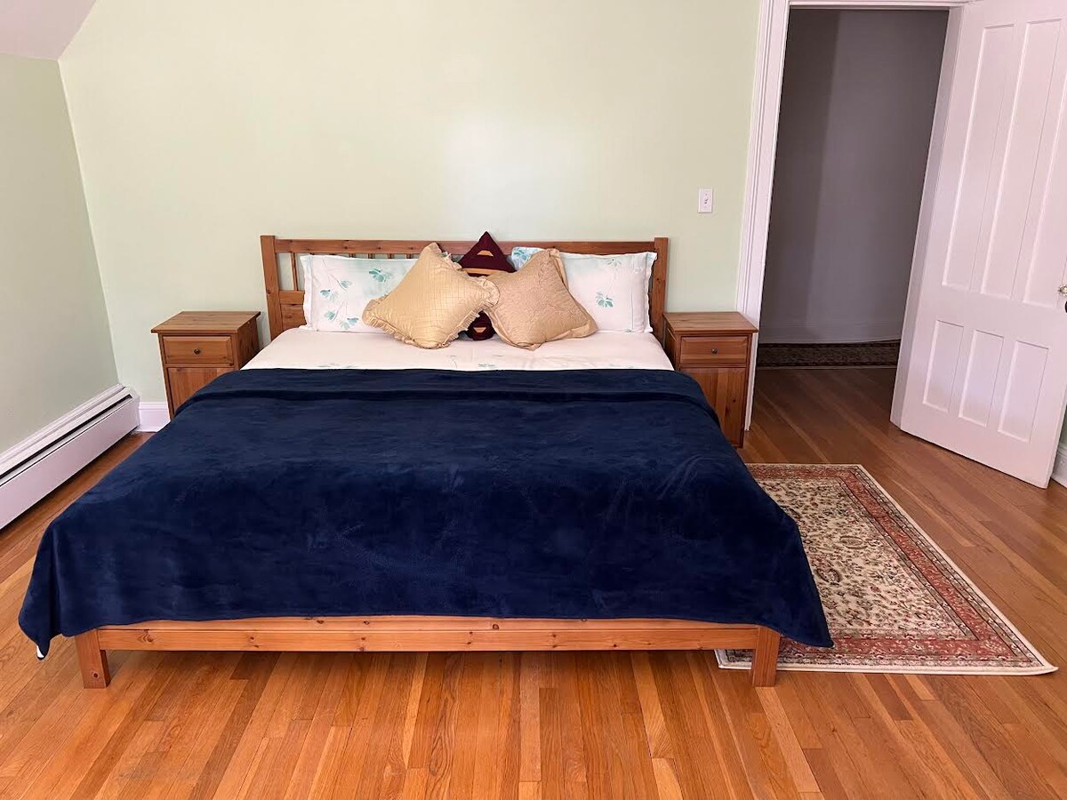A spacious bedroom features a wooden bed frame with a plush navy blanket and decorative pillows. Two nightstands with lamps are positioned beside the bed. Warm hardwood floors are complemented by a small area rug, and a doorway leads to another area of the home.