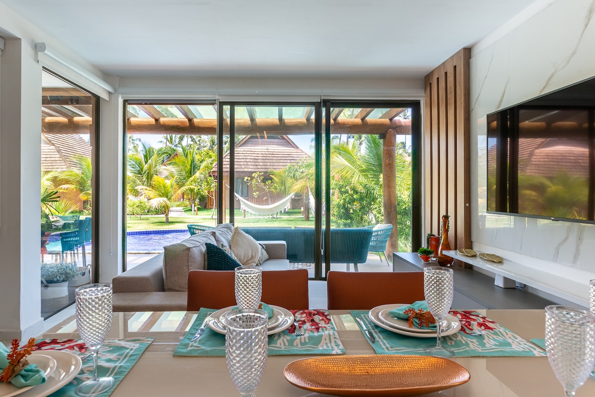 A bright dining area features a modern table set with glassware and decorative plates. Large glass doors open to a lush outdoor view, showcasing a hammock and pool area surrounded by greenery. A cozy seating arrangement with blue upholstery can be seen in the background.