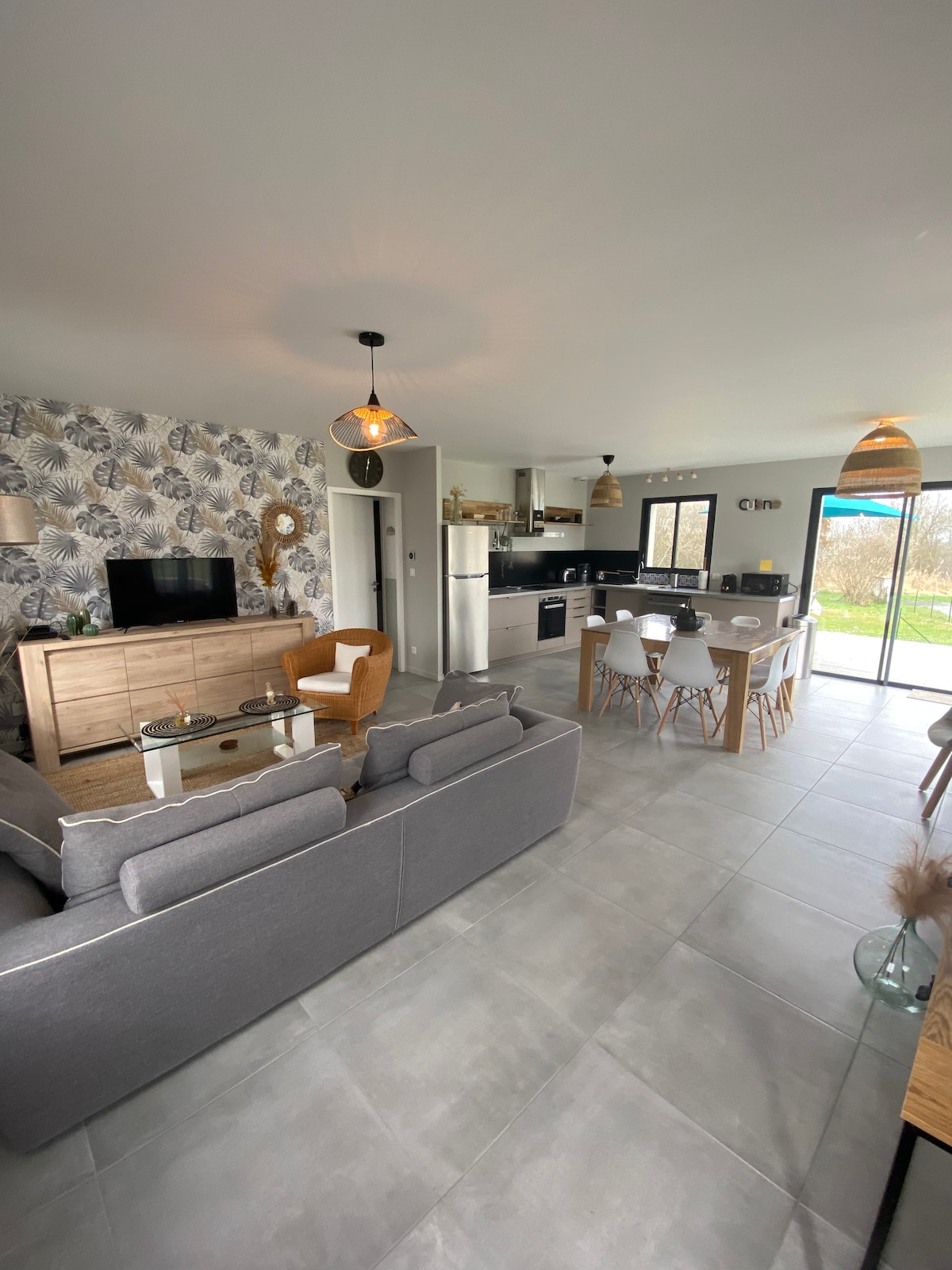 A spacious living area features a modern grey sofa facing a wooden TV cabinet. A dining table with white chairs sits nearby. The open kitchen, equipped with appliances, is visible. Large glass doors lead to an outdoor terrace, allowing natural light into the space.
