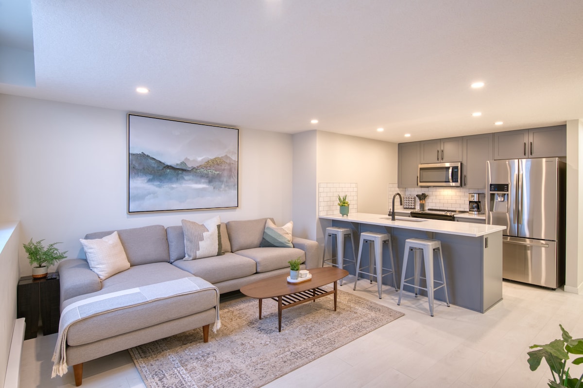 An open-concept living space features a modern kitchen with stainless steel appliances and gray cabinetry. A spacious sectional sofa and a wooden coffee table occupy the living area, while a framed mountain landscape hangs on the wall, adding a subtle touch of decor.