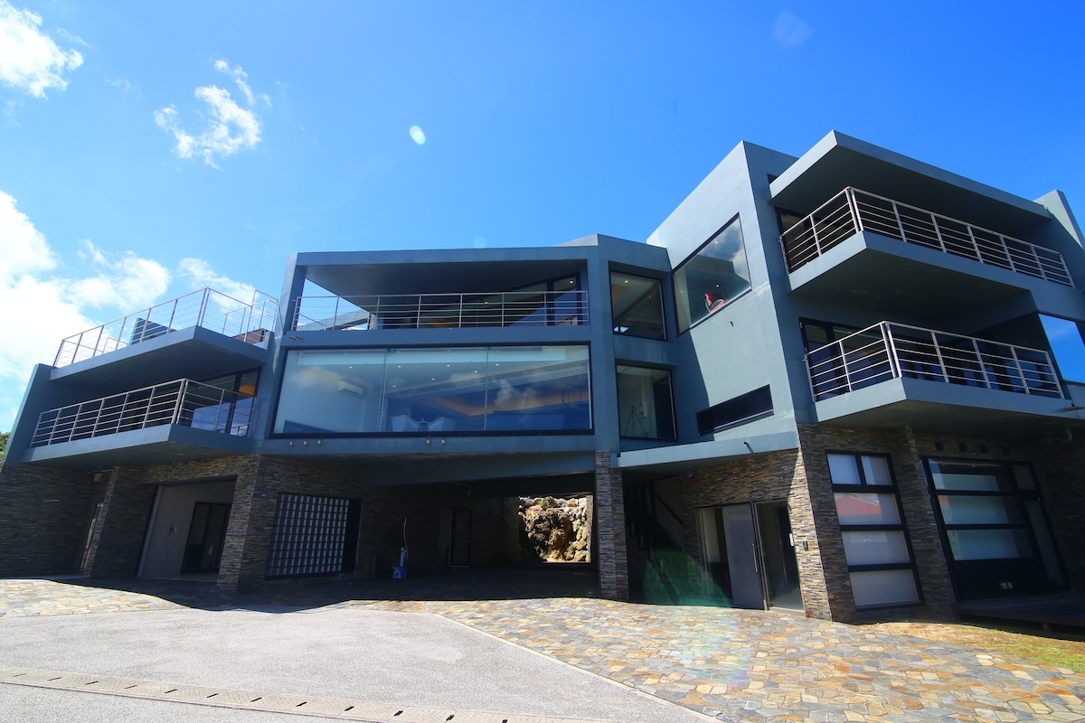 The villa's modern facade is depicted, featuring multiple levels with expansive glass railings. The stone ground is complemented by a structured layout, allowing for architectural depth. Large windows reflect the clear blue sky, enhancing the contemporary design of the property.