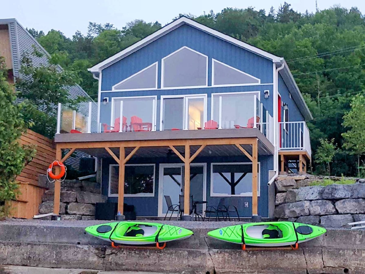 A charming waterfront bungalow is set against a natural backdrop, featuring a walkout basement and a spacious deck. Two green kayaks are docked on the stone shore, while red chairs on the deck provide seating for relaxation, all framed by lush greenery in the distance.