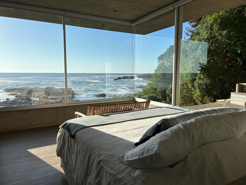 A spacious bedroom is presented with a large window offering an unobstructed ocean view. A wooden bench sits by the window, complemented by a neatly made bed featuring crisp linens, creating a serene and relaxing atmosphere.