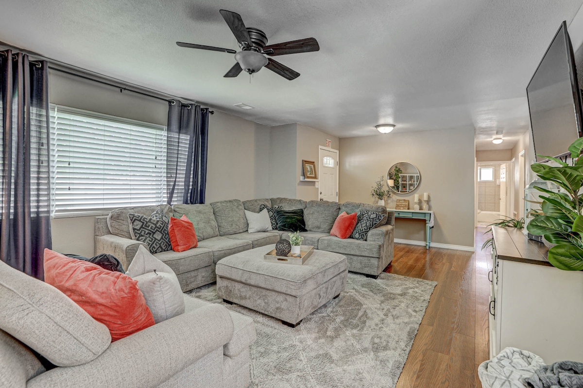 A roomy living area is furnished with a large sectional couch, accented by bright cushions. Light filters through the window, draped with navy curtains. A textured area rug is laid on polished wood flooring, and a ceiling fan enhances the comfortable atmosphere.