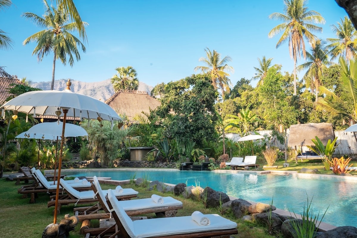 A serene swimming pool is surrounded by tropical greenery and palm trees. Sun loungers are positioned along the poolside, inviting relaxation. Umbrellas provide shaded spots, and a natural landscape adds to the tranquil atmosphere of the resort.