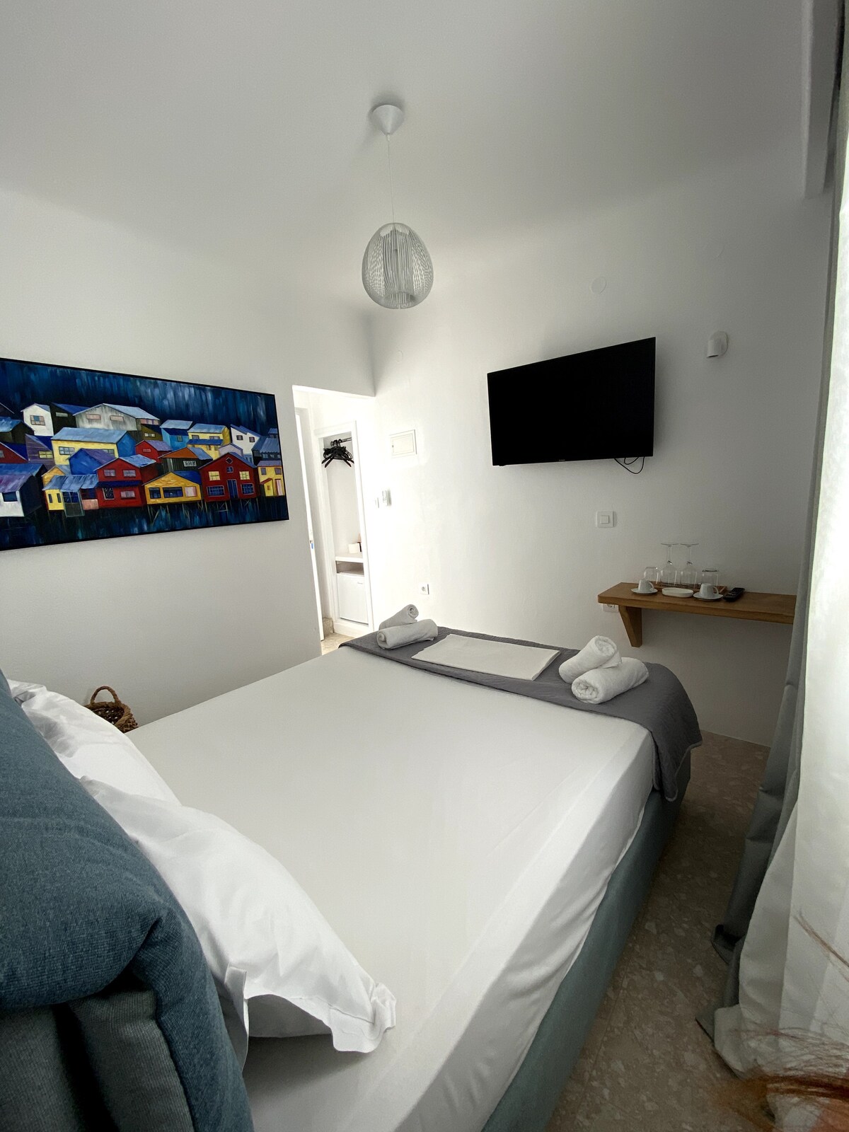A bright guest room is displayed, featuring a spacious bed with soft linens. A piece of colorful wall art is hung above the bed, while a television is mounted on the wall. A small table with glasses is placed nearby, and a door leads to an adjoining area.