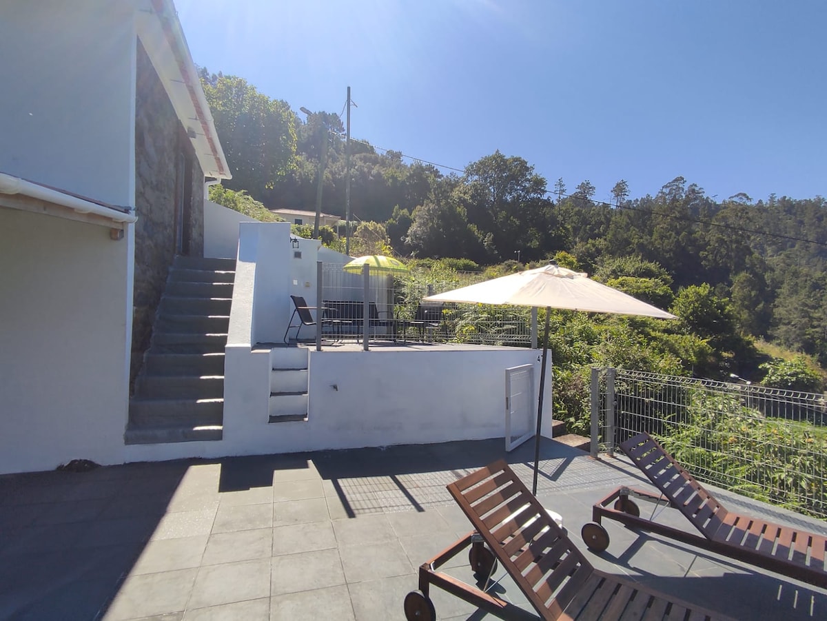 The area features a spacious terrace with lounge chairs and umbrellas, surrounded by green vegetation. Steps lead from the terrace to the entrance of the villa. Clear blue skies enhance the open and inviting outdoor space.