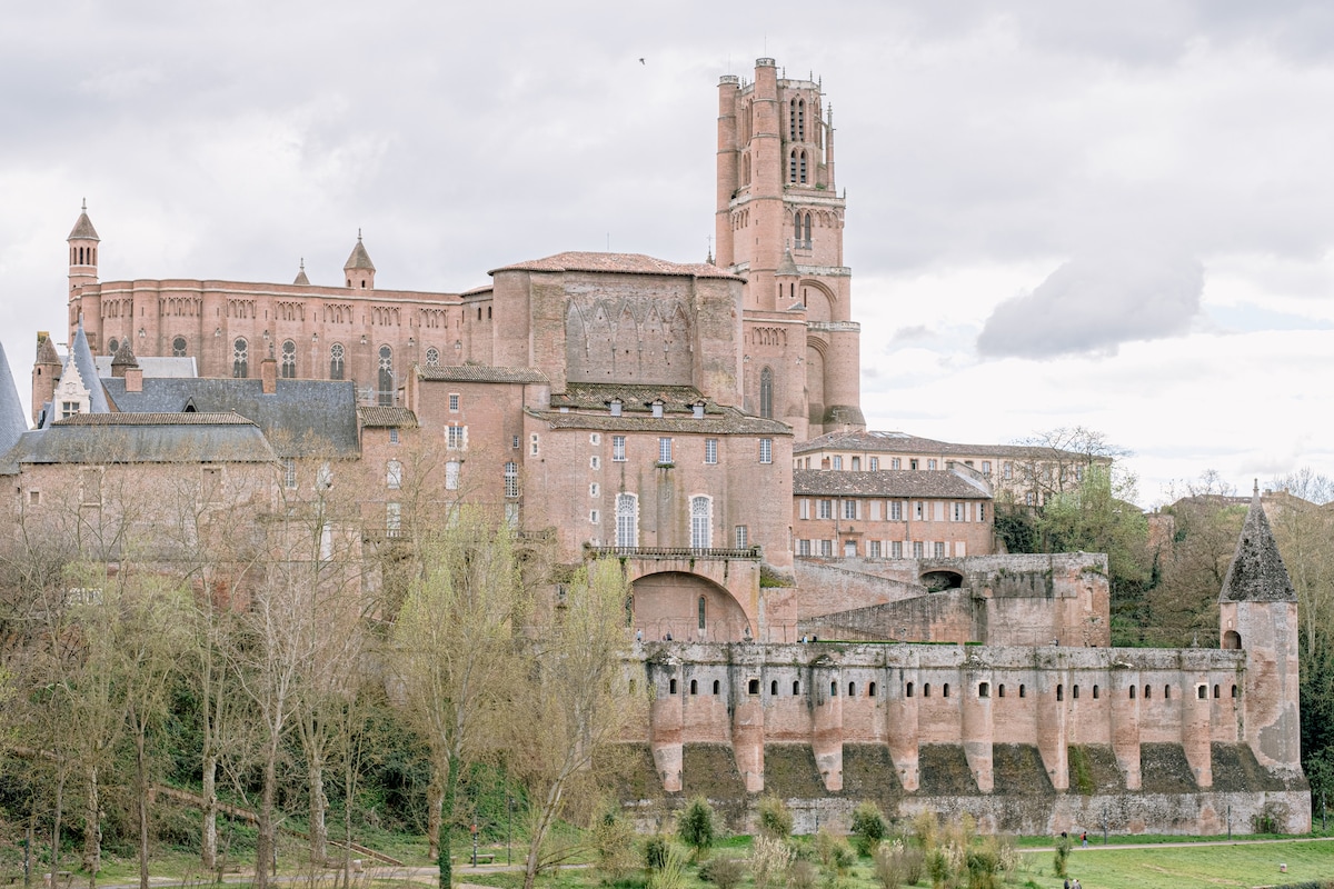 A historic building complex is visible, featuring stone structures with varying heights and architectural details. Tall towers rise above, surrounded by trees and lush greenery. The view includes a bridge and a section of water reflecting the surroundings, creating a serene landscape.