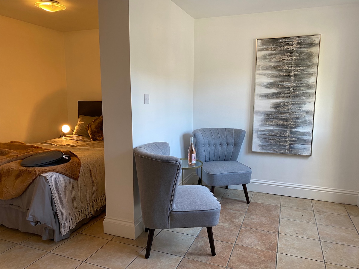 A cozy corner of the studio features two grey chairs positioned beside a small table. The chair upholstery is plush, and a bottle of wine rests on the table. In the background, a spacious bed is visible, draped with a textured blanket.