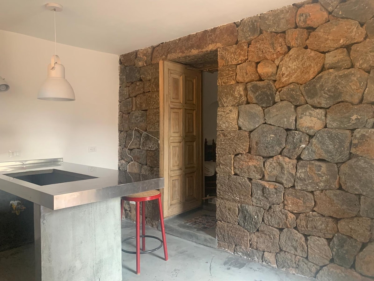 The kitchen area showcases a minimalist design with a concrete countertop and a red stool. A rustic stone wall is featured prominently, complementing the warm wooden door that leads to the bedroom. Soft lighting is provided by an overhead pendant lamp.
