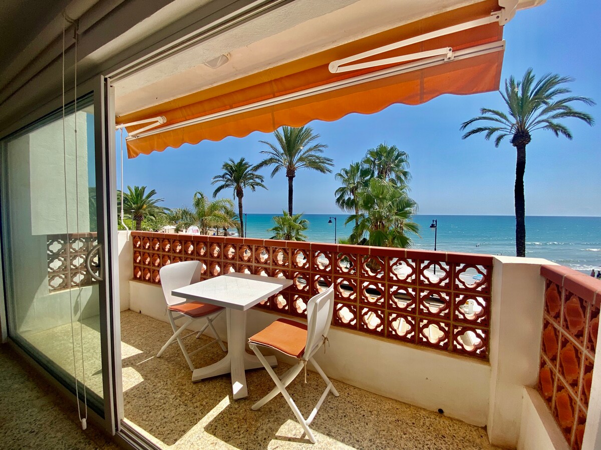 A bright balcony features a small table and two chairs, offering views of palm trees and the ocean. The space is shaded by an orange awning, enhancing the outdoor experience. A nearby glass door provides access to the interior, allowing natural light to flow in.