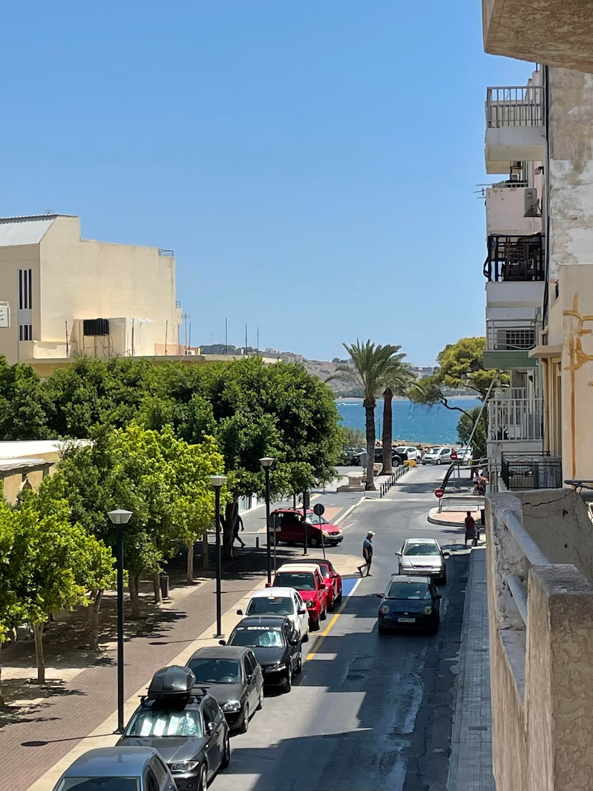 A view from the balcony reveals a quiet street lined with parked cars. Lush trees provide shade along the walkway, with the glimmering blue sea visible in the distance. Palm trees can be seen swaying gently in the breeze, enhancing the coastal ambiance.