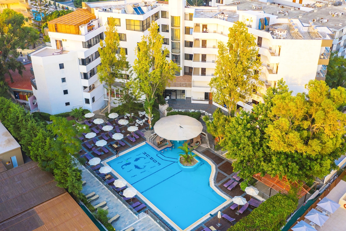 An aerial view showcases the spacious pool surrounded by umbrellas and lounge chairs. The elegantly designed building features multiple stories with large windows, complementing the greenery of trees and shrubs around the pool area.