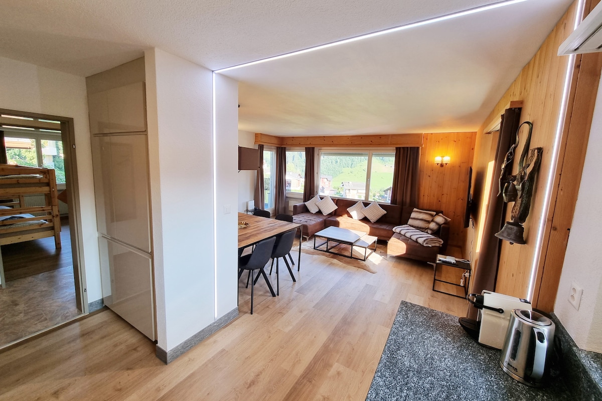 An open living and dining area is showcased, featuring a modern dining table surrounded by chairs. A comfortable seating arrangement with cushions is situated beside large windows that provide natural light. The space is adorned with light wood finishes, creating a warm atmosphere.