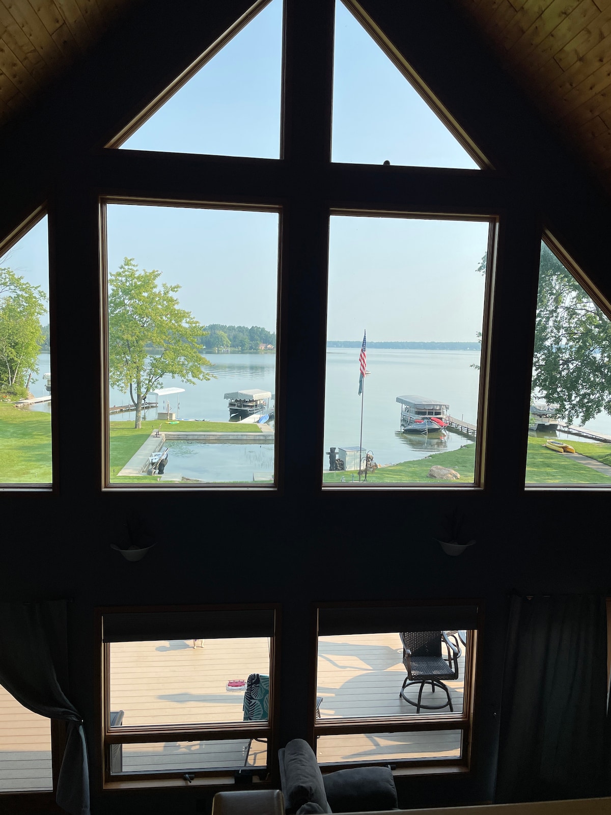 A large open window offers a panoramic view of Long Lake, framed by the triangular architecture of the home. The scene includes boats tied to the dock, a grassy yard, and trees lining the shore, contributing to the serene lakeside setting.