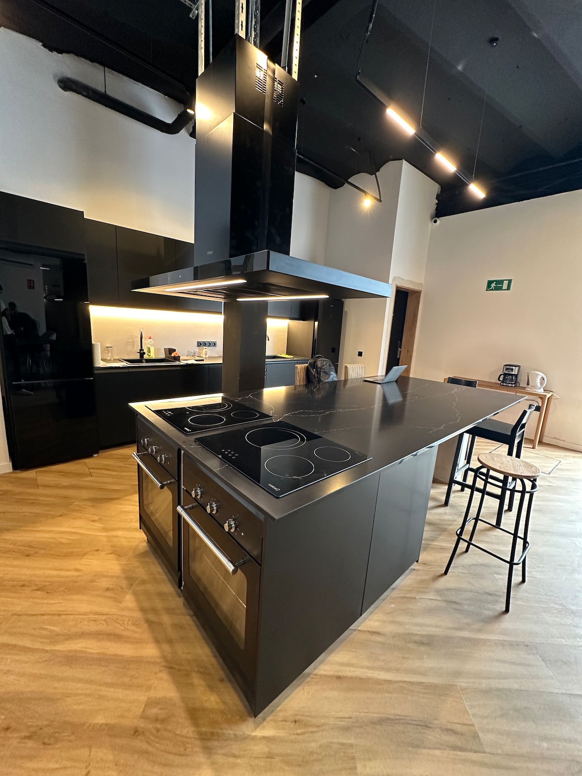 A modern kitchen design features sleek black cabinetry and an island equipped with a four-burner cooktop. Stainless steel appliances, including an oven, are integrated into the cabinetry. Additional seating is provided by two high stools at the island, illuminated by overhead lighting.