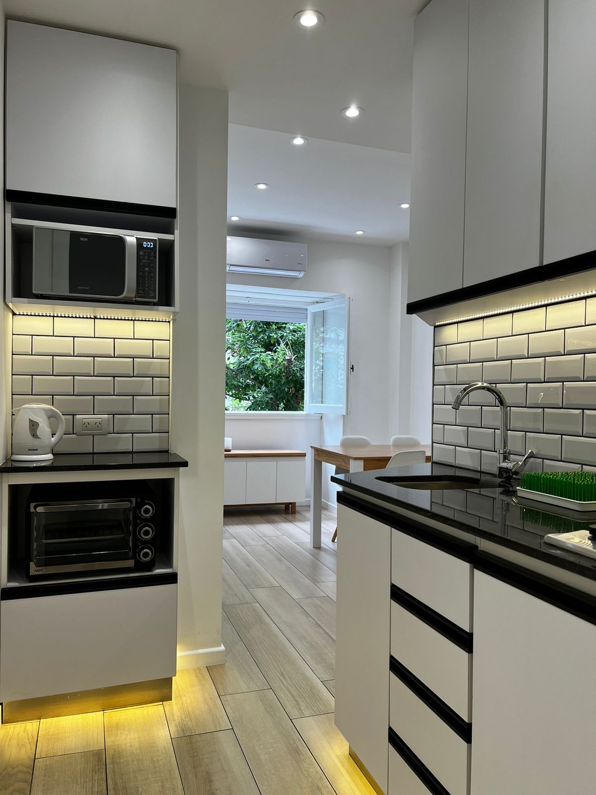 A modern kitchen is presented with sleek cabinetry and a black countertop. Under-cabinet lighting highlights the textured backsplash. A microwave and oven are visible, with a kettle on the counter. A window opens to a view of greenery, suggesting a bright and airy atmosphere.