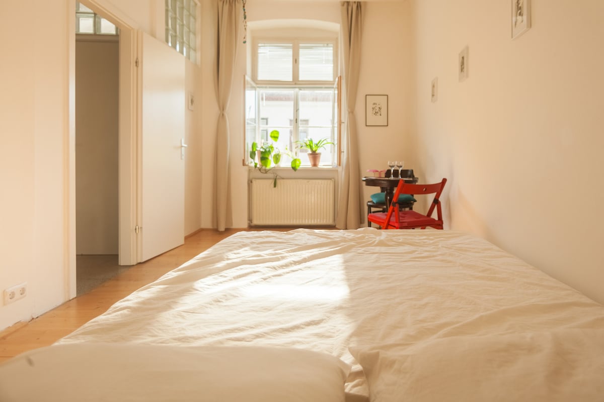A serene sleeping area features a spacious bed with soft linens. Natural light enters through a large window, illuminating a small table adorned with plants. A red chair adds a pop of color against the neutral-toned walls.