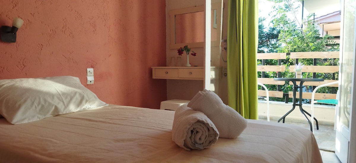 A cozy bedroom features a spacious bed with white linens and neatly rolled towels placed on top. Light enters through an open door leading to a balcony with a small table and chairs, surrounded by greenery.