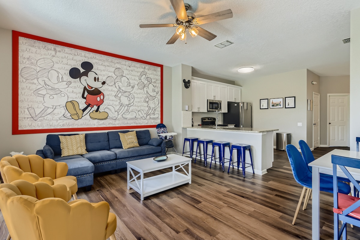The spacious living area features a large blue sectional sofa facing a coffee table. A vibrant mural of Mickey Mouse adorns the wall, complemented by yellow armchairs and blue barstools at the kitchen counter. The open kitchen area includes modern appliances and bright cabinetry.