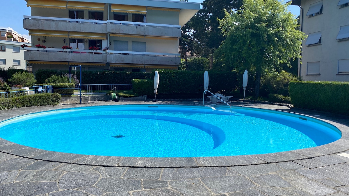 A centrally located pool is surrounded by a neat stone walkway and well-maintained gardens. Sun loungers are positioned around the edge of the water, while vibrant greenery provides a tranquil setting. The pool features a circular design with clear, inviting blue water.