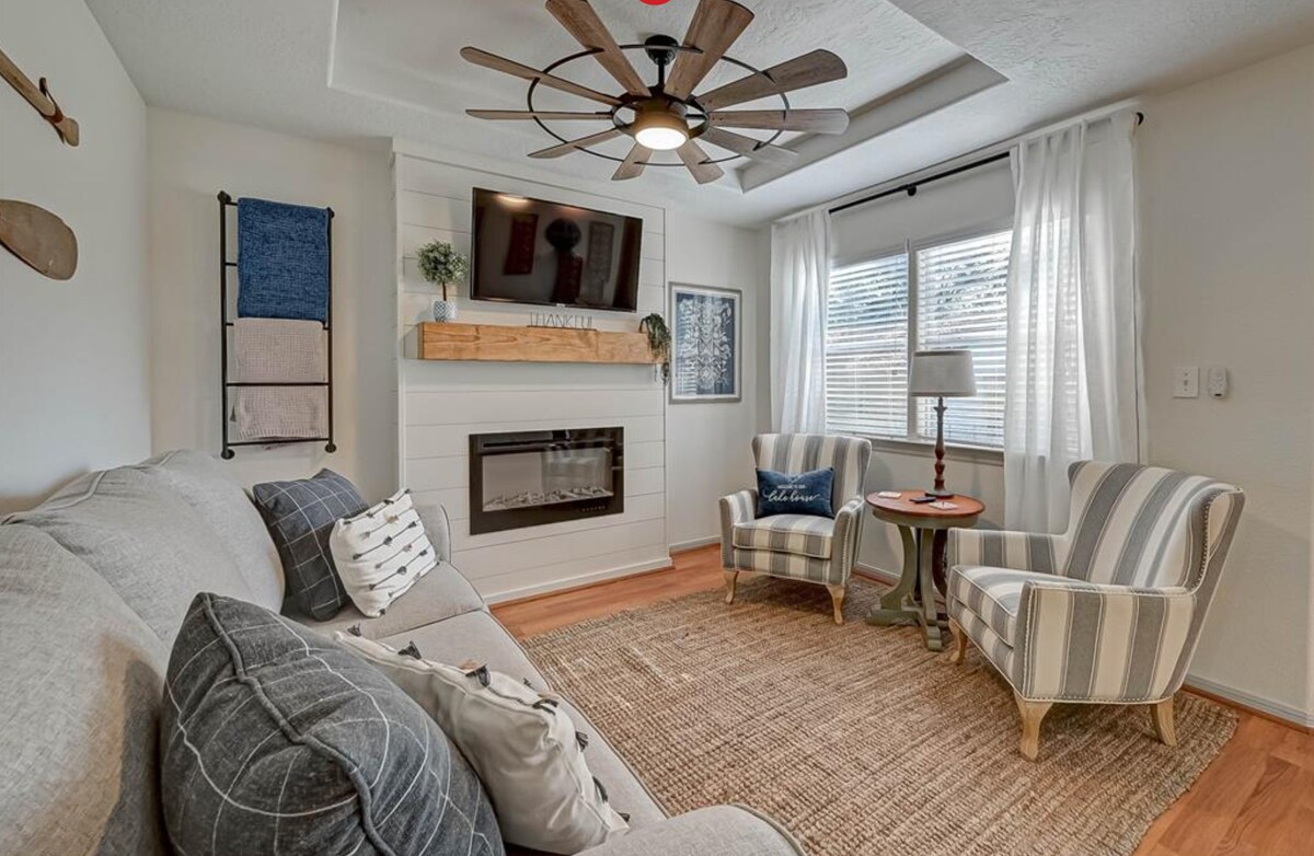 A comfortable living area features a light-colored sofa with decorative pillows. Two patterned chairs are positioned beside a stylish coffee table. Natural light filters through the windows adorned with sheer curtains, while a modern ceiling fan adds character above. A decorative fireplace adds warmth to the space.
