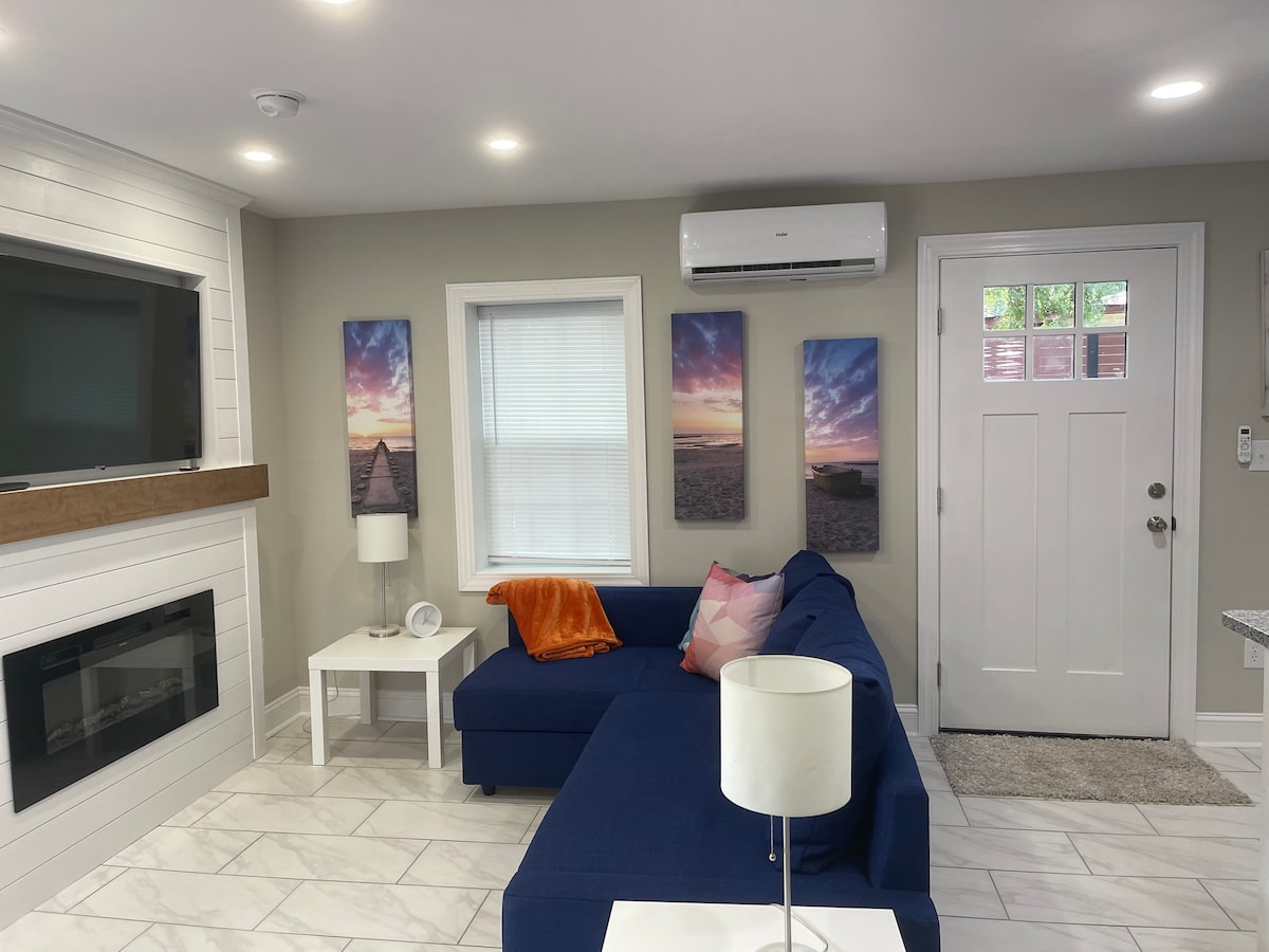 A modern living area features a spacious blue sectional sofa and a flat screen TV mounted above a stylish electric fireplace. Light-colored walls are enhanced by photographs of sunset landscapes. A door leads to the exterior, with natural light filtering through the window.