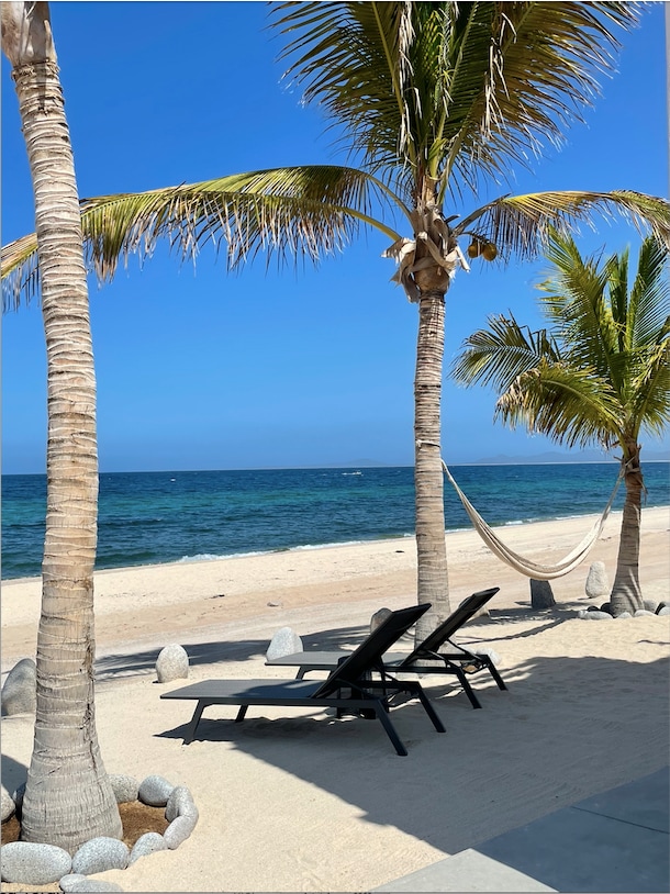 Two lounge chairs are positioned on the sandy beach, framed by palm trees swaying in the gentle breeze. A hammock hangs invitingly between the trees, overlooking the clear ocean waters under a bright blue sky.