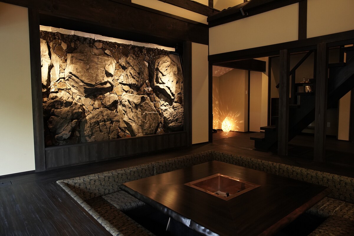 A spacious indoor area is highlighted by a striking rock wall, softly illuminated to enhance the natural texture. A low wooden table with a built-in fire pit serves as a central gathering point. Subtle lighting casts gentle shadows, creating a warm and tranquil ambiance.