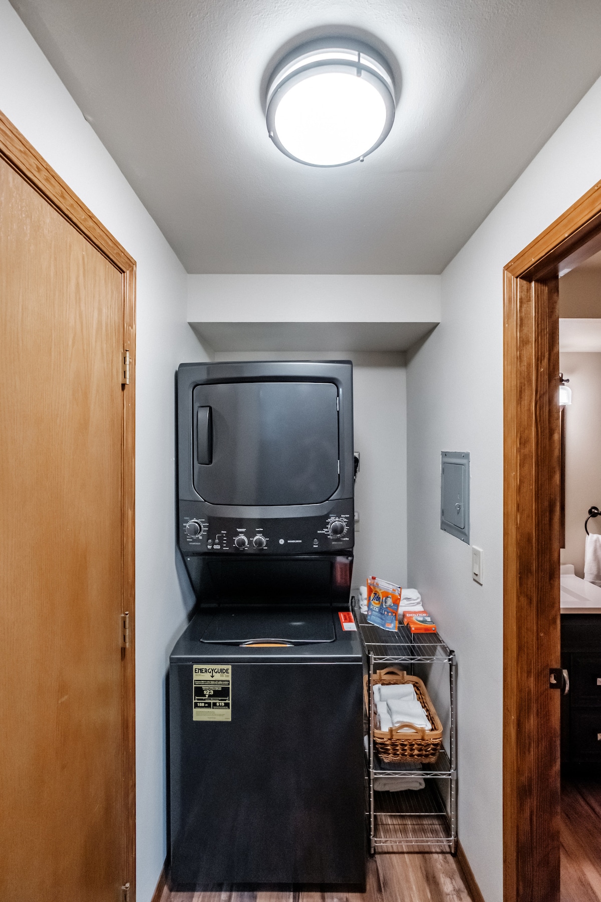 A compact laundry area is equipped with a stacked washer and dryer unit, featuring a simple control panel. A small cart holds laundry supplies, while a light fixture above provides illumination.