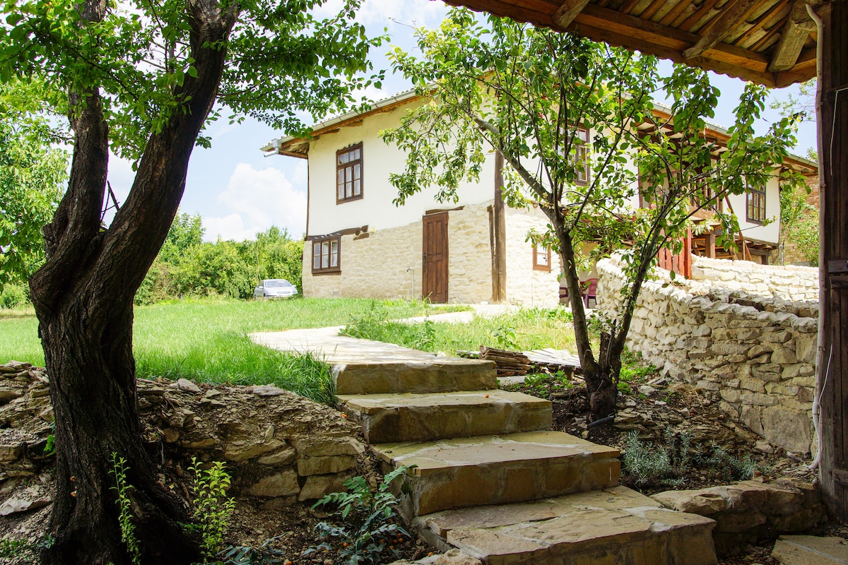 A traditional Bulgarian house is nestled among greenery, with a stone pathway leading to its entrance. The building features wooden windows and a light-colored facade, surrounded by lush grass and trees. The outdoor space offers a serene environment for relaxation.