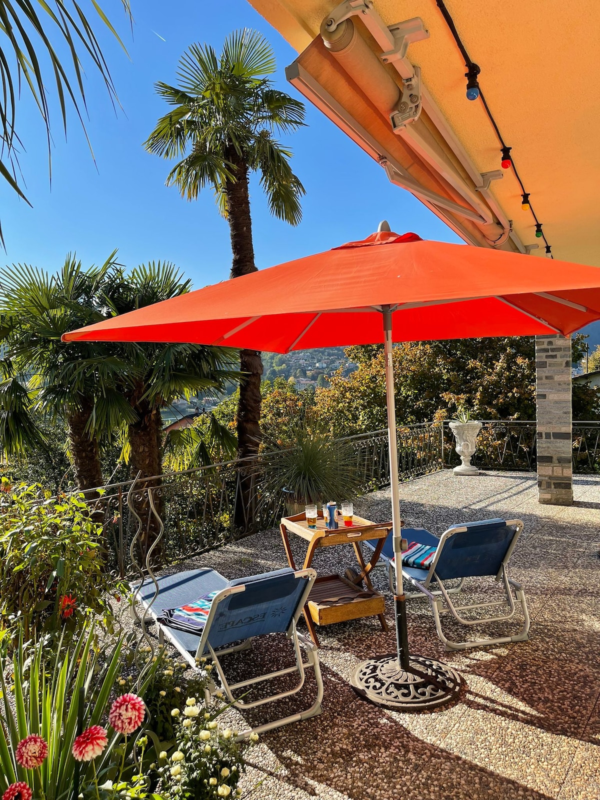 A large terrace is featured with an orange umbrella providing shade over a small table and two chairs. Tropical plants flank the area, and a glimpse of the landscape and distant mountains can be seen against a clear blue sky.
