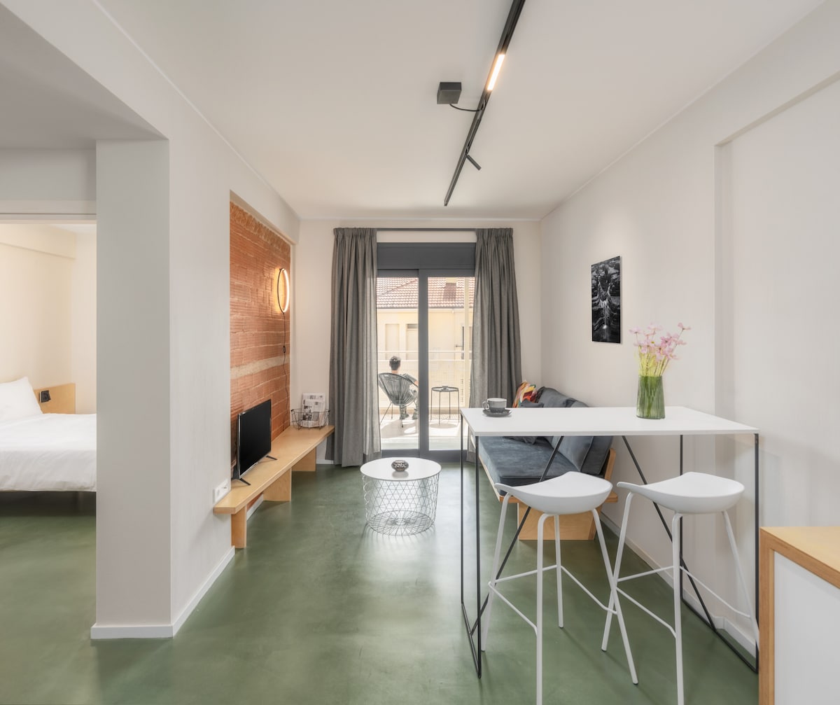 A modern living space features a dining table with white stools, complemented by a grey sofa. Natural light fills the room, illuminating minimalist décor. A glass door leads to a balcony, while a television is positioned on a wooden shelf against a textured wall.