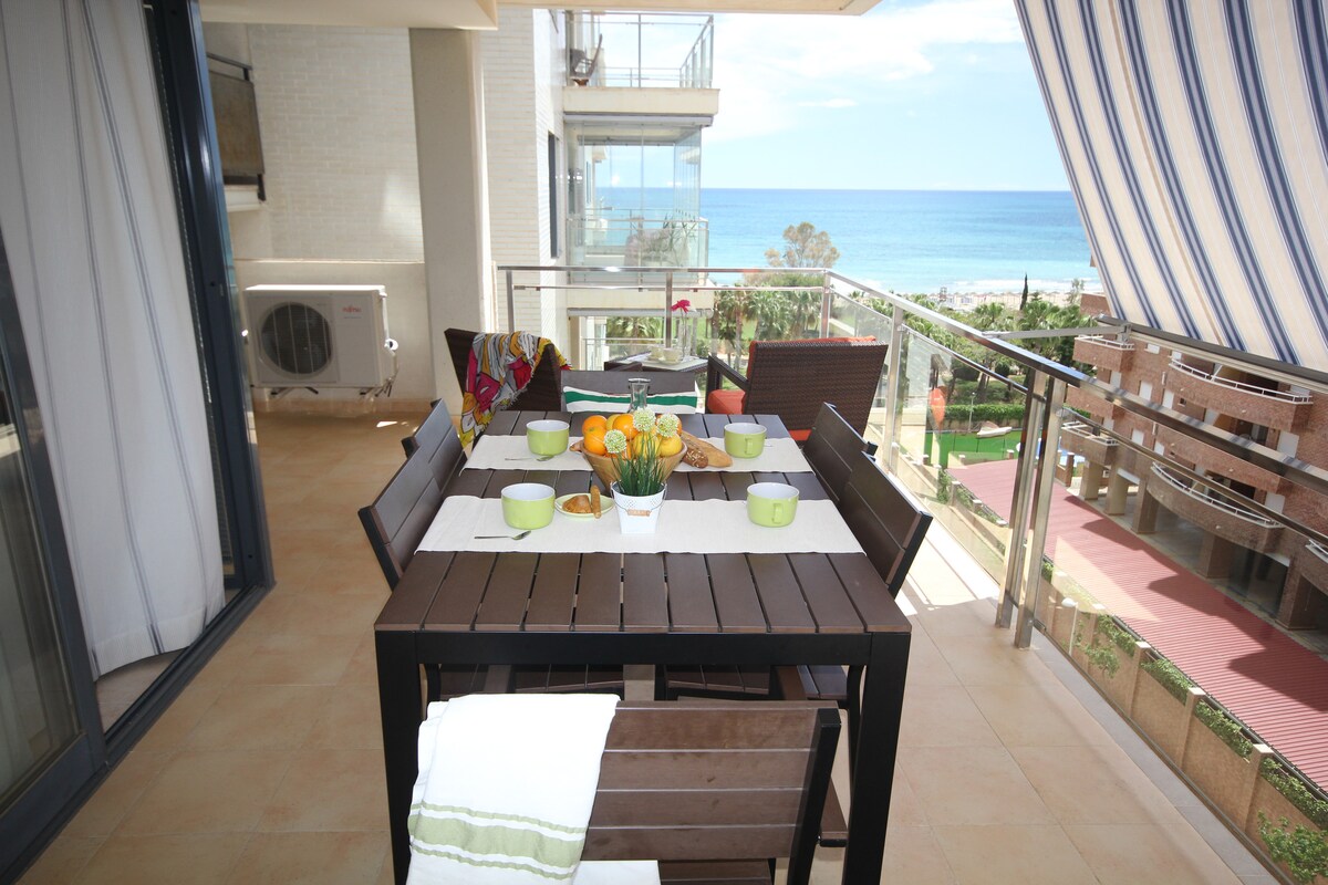 An outdoor terrace features a wooden dining table set for six, adorned with vibrant bowls of fruit and cups. The view overlooks the tranquil sea and well-maintained green areas. A soft breeze is suggested by the partially closed striped awning above.