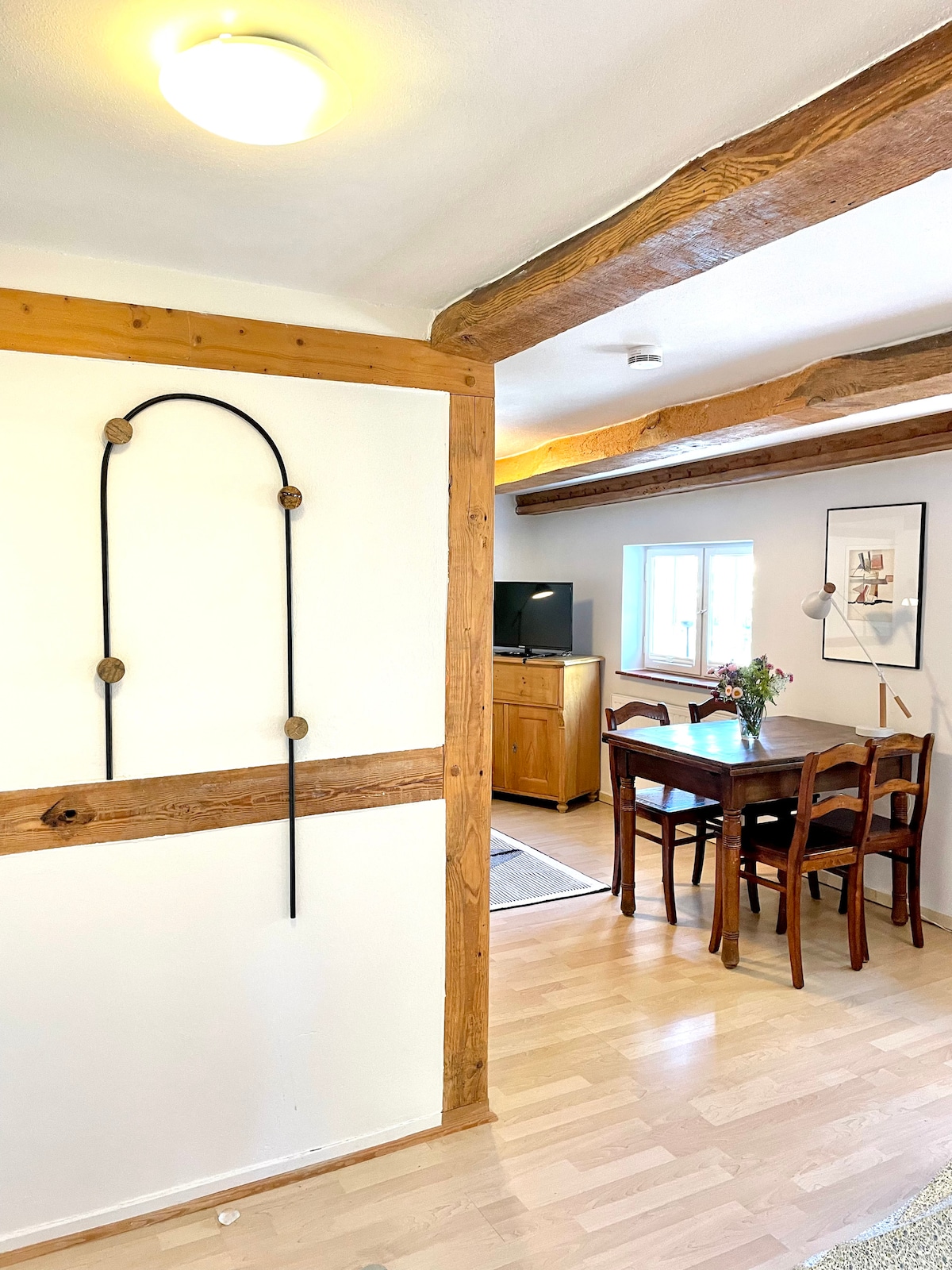 A warm entryway features wooden beams and a decorative wall piece, leading to a dining area. A rectangular table with wooden chairs is positioned under natural light from a nearby window, complemented by a sideboard and a television in the background.