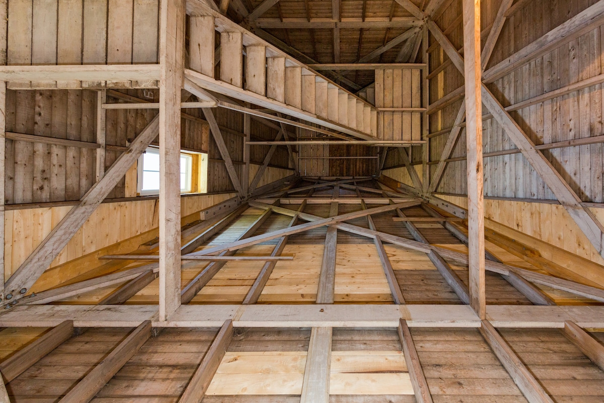 The interior structure of the lighthouse showcases wooden beams and an open ceiling, emphasizing the height of the tower. Stairs ascend along one side, leading to upper levels. A small window allows natural light to enter, enhancing the wooden aesthetics.