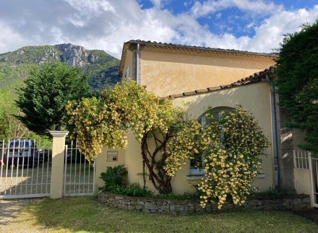 The exterior of a charming house is framed by lush greenery and a flowering vine. A stone wall borders the garden, while a driveway leads to a gated entrance. A mountainous landscape rises in the background, enhancing the natural setting.
