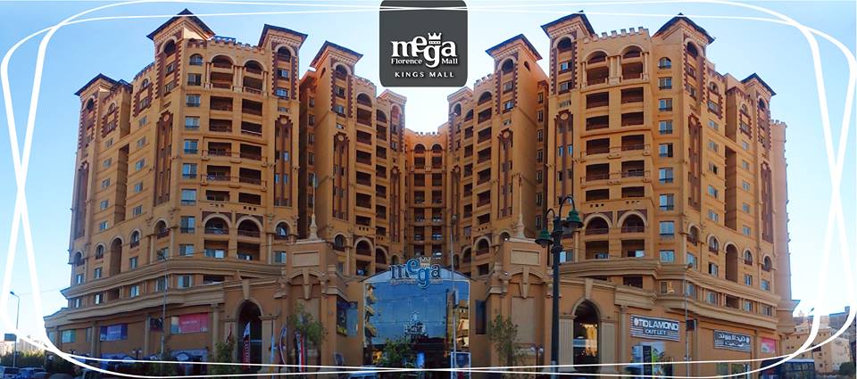 A large modern building with an intricate architectural design is showcased. The facade features warm tones and decorative elements, framed by two symmetrical towers. Glass entrances and storefronts are presented at the ground level, providing a view of the commercial activity within.