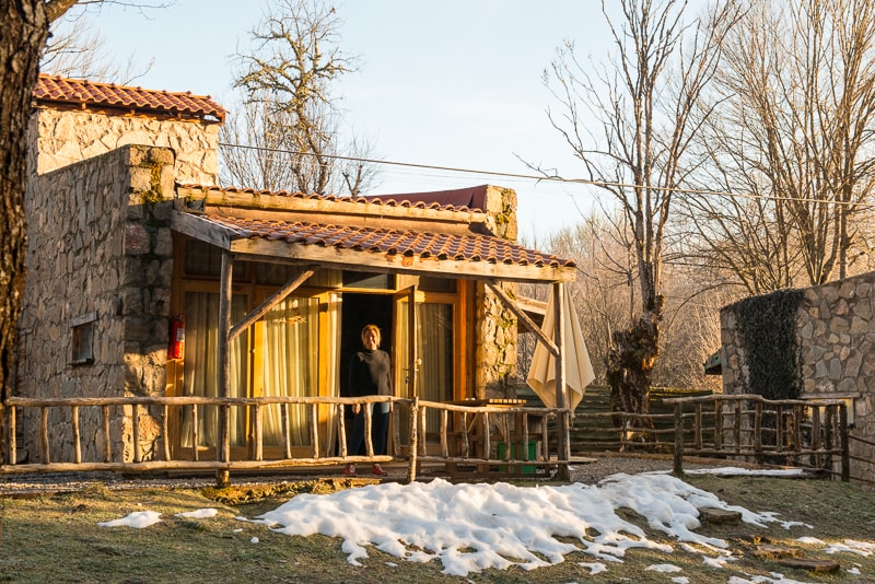 A charming stone cottage is highlighted in this image, featuring a wooden porch with a gently sloped roof. Large windows allow natural light to fill the interior, while a small patch of snow is visible on the ground nearby. Leafless trees frame the scene, adding to the serene atmosphere.