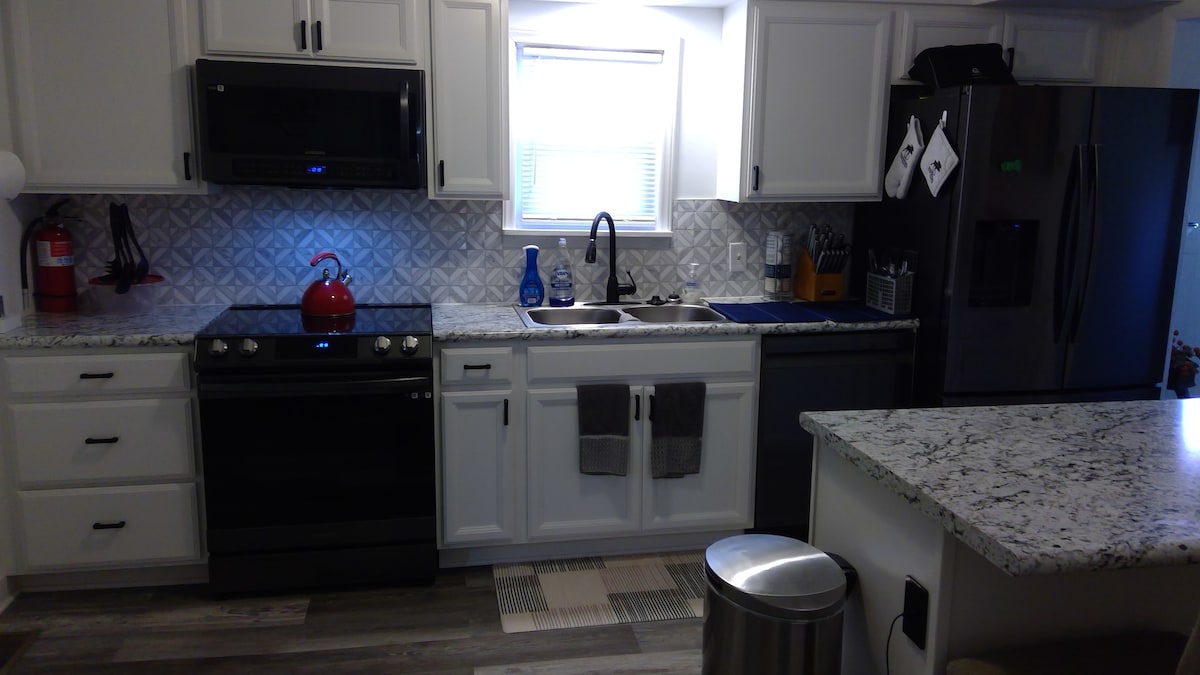 The kitchen features modern white cabinetry with granite countertops, and a black stove and microwave are positioned above one another. A sink is located under a window, surrounded by utensils and a blue dish rack. A stainless steel refrigerator and trash can are visible.
