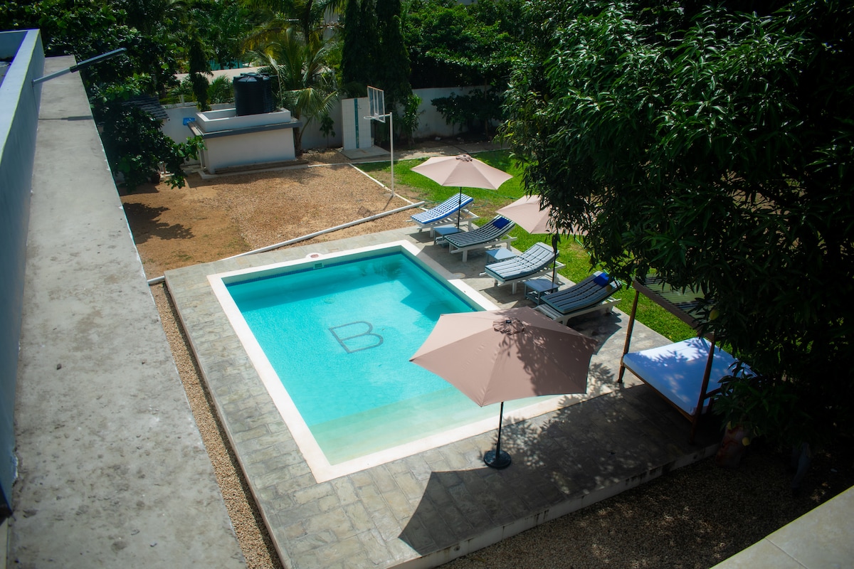 A serene outdoor area features a clear swimming pool surrounded by sun loungers and shaded umbrellas. Lush greenery and well-maintained garden spaces enhance the tranquil atmosphere, providing a relaxing environment for guests. The pool area is designed for leisure and enjoyment.