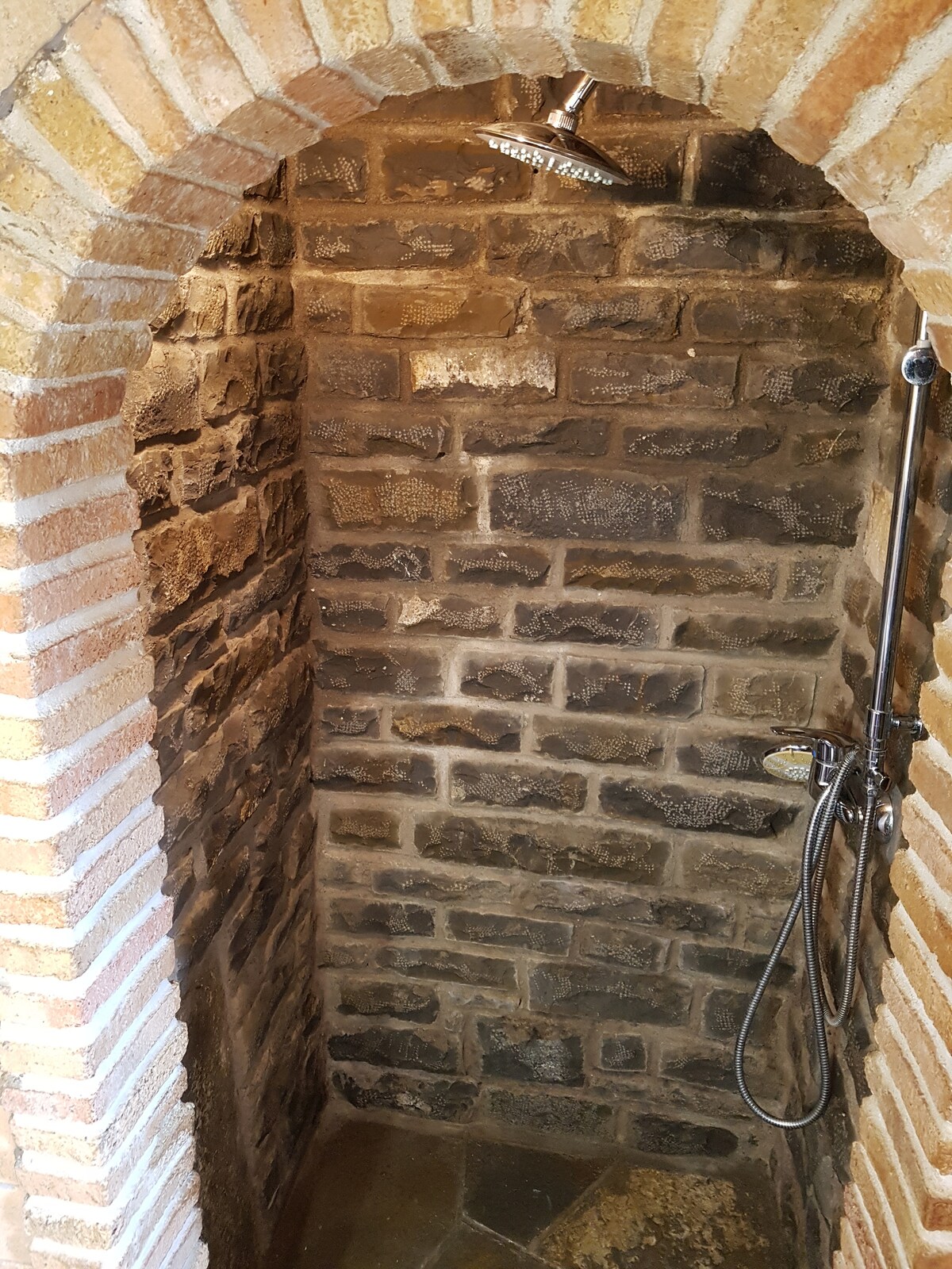 An enclosed shower area features rugged stone walls, enhancing the rustic charm. A rainfall showerhead is mounted overhead, while a handheld shower fixture is attached to the wall. The stone flooring adds to the natural aesthetic of the space.