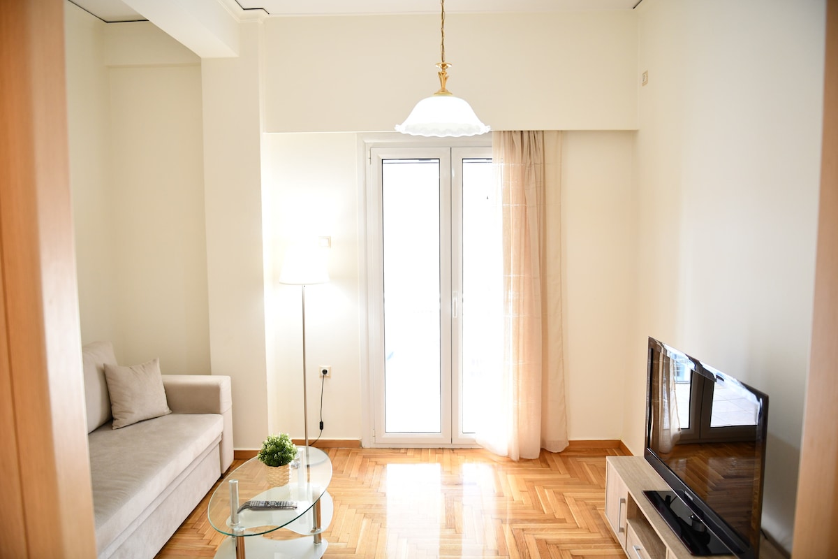 The living room showcases a light, airy ambiance with large glass doors allowing natural light to fill the space. A comfortable sofa sits adjacent to a coffee table, while a TV is positioned on a cabinet, adding functionality to the area.
