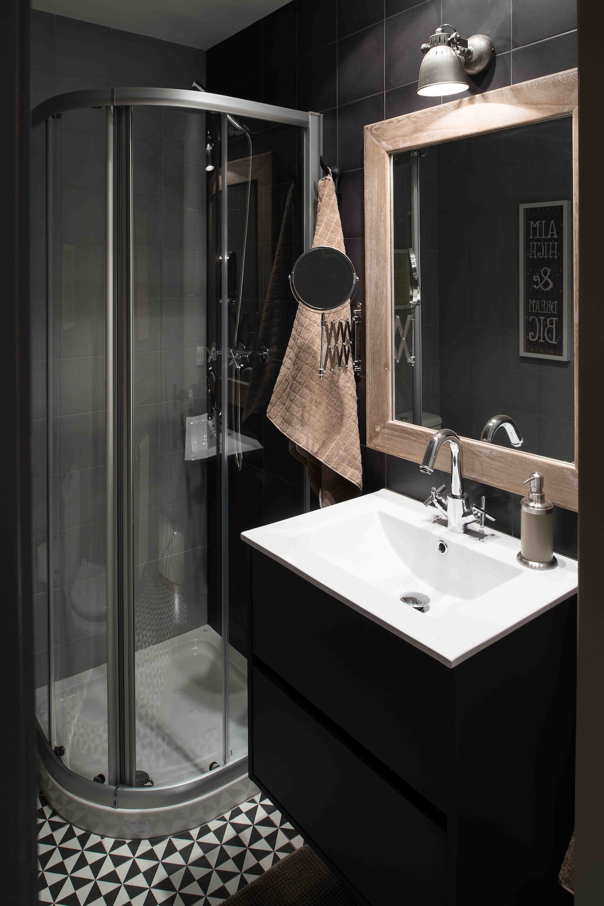 A compact bathroom features a glass shower cabin alongside a modern, white sink set on a sleek, dark cabinet. A large mirror framed in natural wood complements the stylish decor. Decorative tiles add texture to the floor.